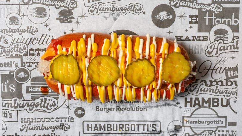 A Hamburgotti's hot dog with mustard, mayonnaise, ketchup, and pickles on branded paper.