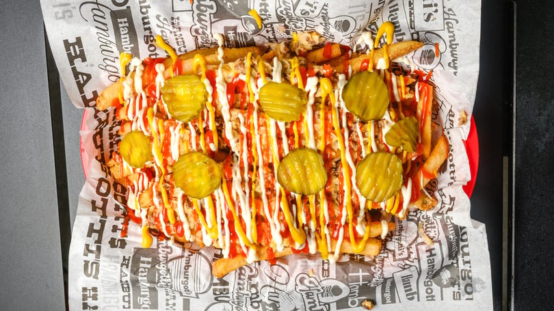 Loaded fries with ketchup, mayonnaise, mustard, and pickle slices in a red basket.